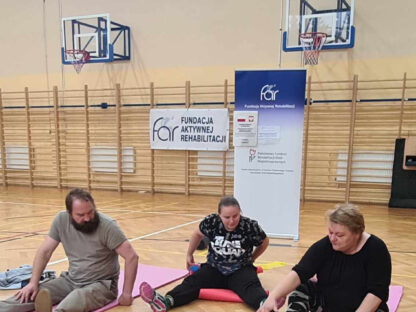 zdjęcie - WAZ Czudec, ćwiczenia z piłką w pozycji siedzącej na matecu na sali gimnastycznej. Ćwiczą trzy osoby.