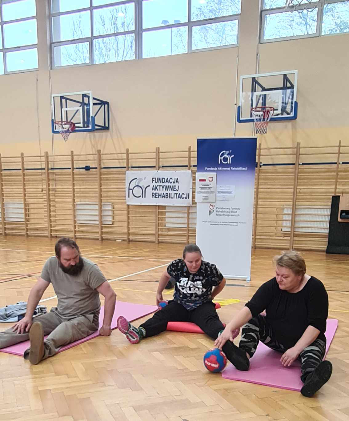 zdjęcie - WAZ Czudec, ćwiczenia z piłką w pozycji siedzącej na matecu na sali gimnastycznej. Ćwiczą trzy osoby.