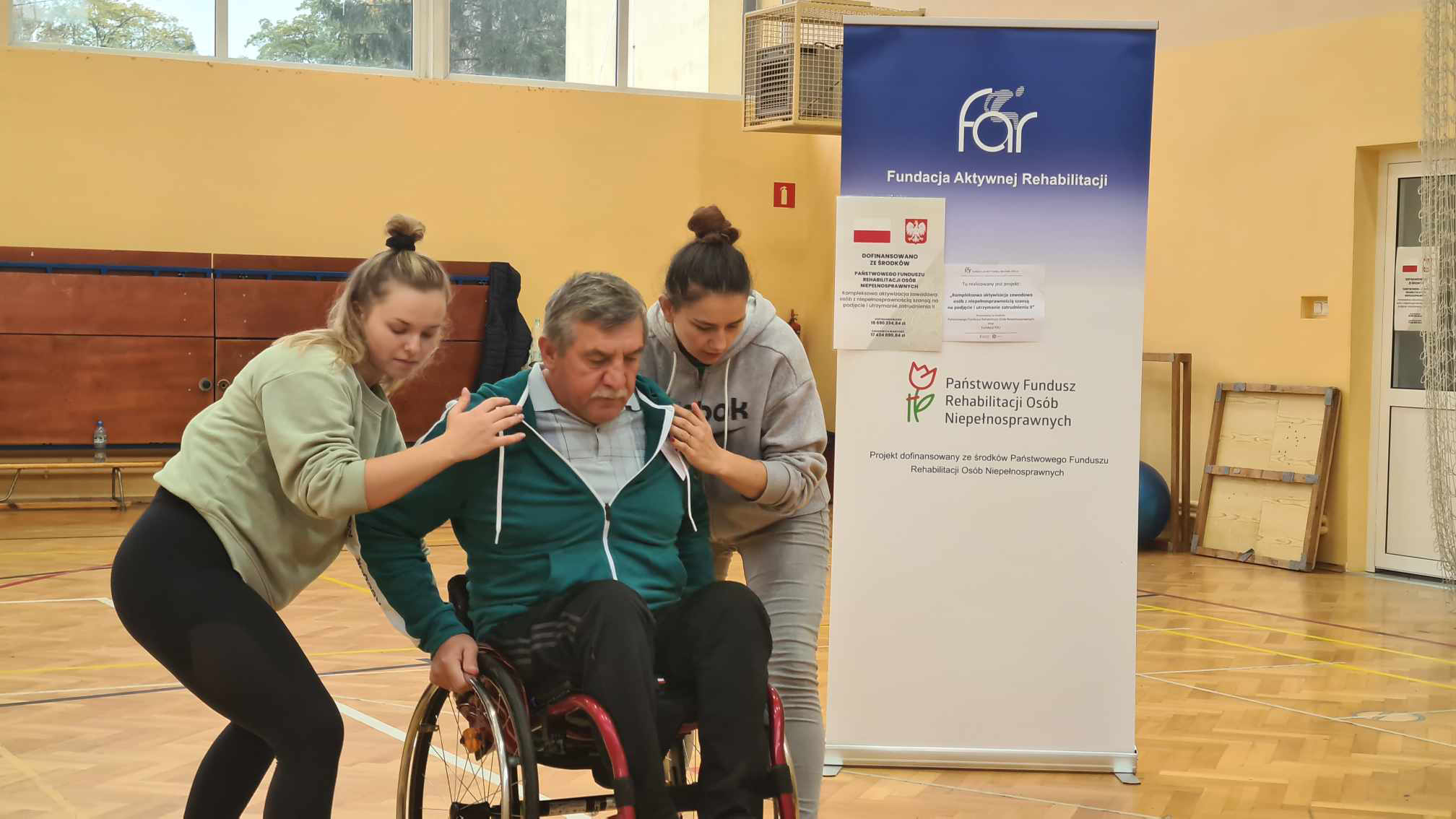WAZ w Czudcu, ćwiczenia na sali. Na zdjęciu uczestnik warsztatu poruszający się na wózku inwalidzkim, asekurowany przez dwie osoby, uczy się przejeżdżać przez progi.