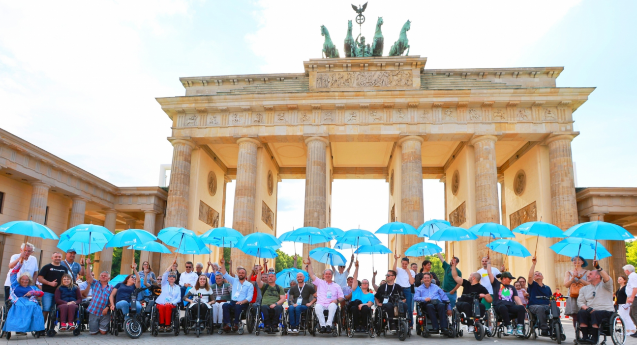 Zdjęcie uczestników kongresu ESCIF w Berlinie. Wszyscy unoszą nad głowami otwarte jasnoniebieskie parasolki, w tle Brama Brandenburska. Kongres ESCIF, 22-25.05.2024
