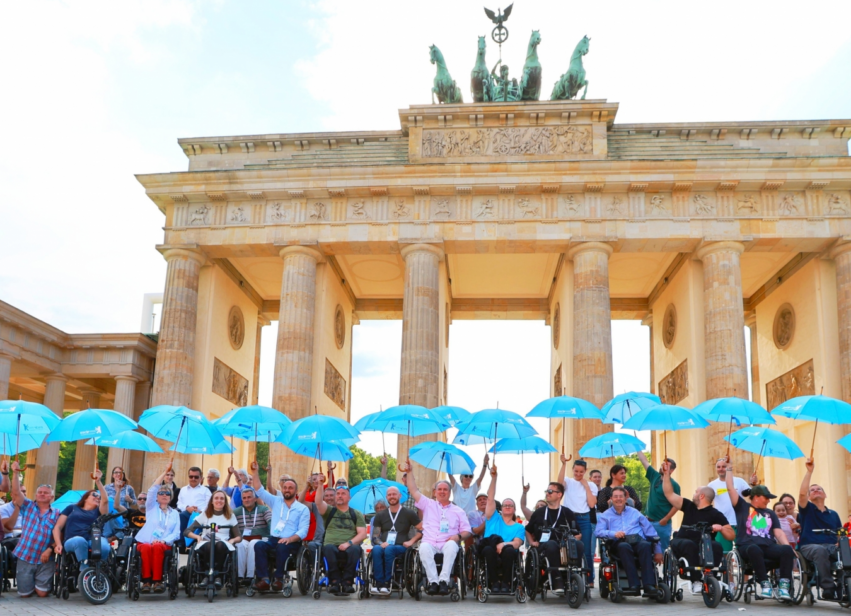 Zdjęcie uczestników kongresu ESCIF w Berlinie. Wszyscy unoszą nad głowami otwarte jasnoniebieskie parasolki, w tle Brama Brandenburska. Kongres ESCIF, 22-25.05.2024