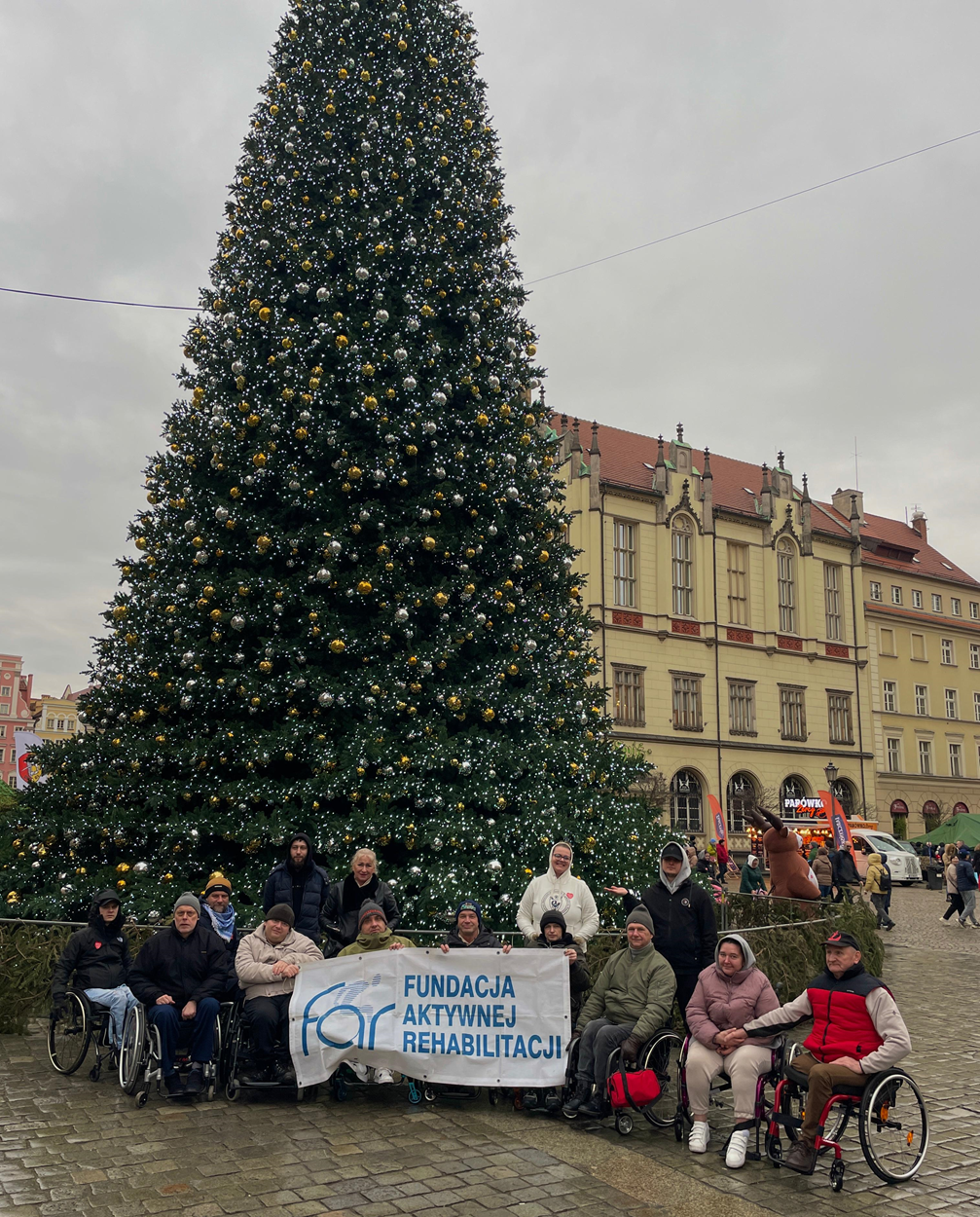 Uczestnicy i kadra obozu z bannerem FAR na rynku w Wrocławiu. W tle ogromna, udekorowana świątecznie choinka. Obóz Aktywnej Rehabilitacji we Wrocław 20-31.01.2025 r.