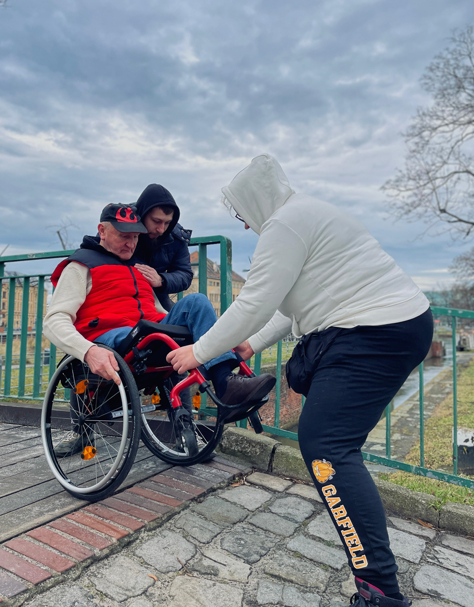 Uczestnik obozu, asekurowany przez dwie osoby, ćwiczy technikę jazdy na wózku w terenie, Obóz Aktywnej Rehabilitacji, Wrocław 20-31.01.2025 r.