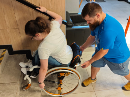 Wolontariusz asekuruje zjeżdżającą po schodach osobę na wózku.