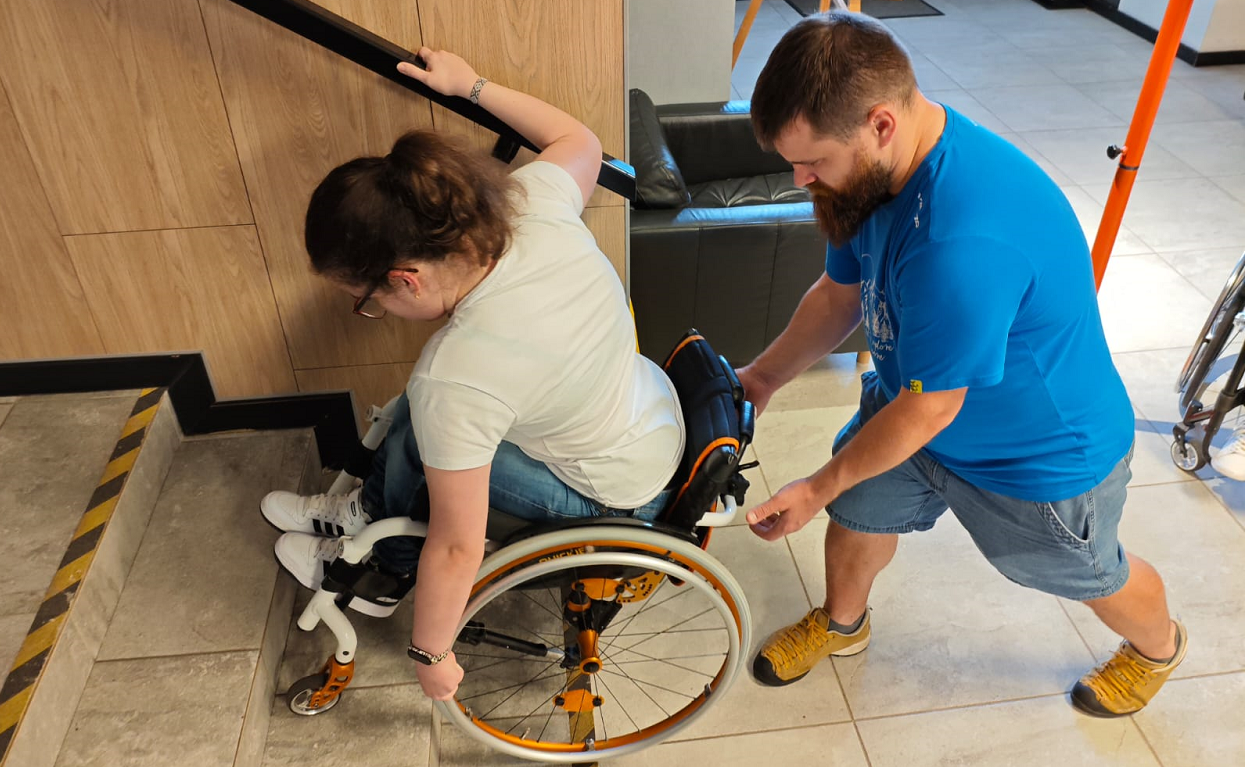 Wolontariusz asekuruje zjeżdżającą po schodach osobę na wózku.